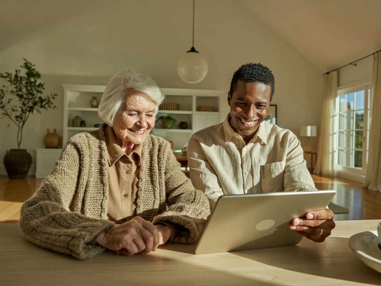 Una signora e una giovane tutor sorridono insieme davanti a un tablet, in una stanza luminosa